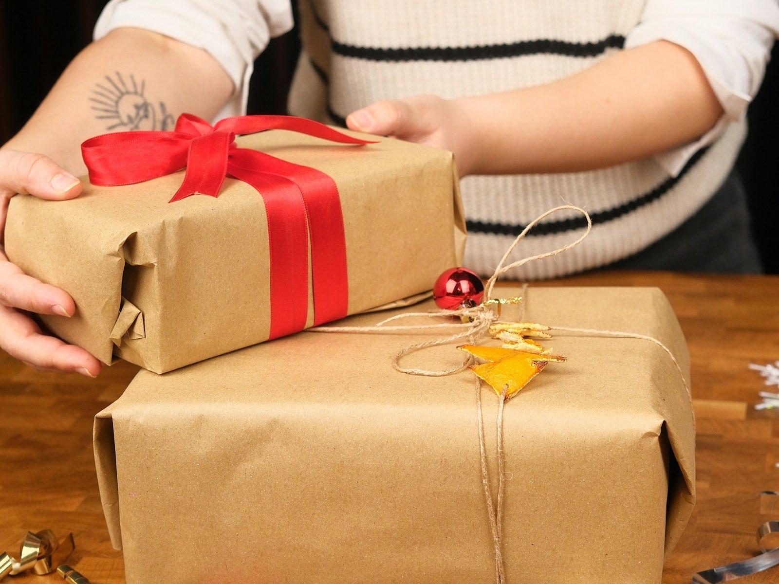 Two hands pressing kraft paper flat around a boxed gift, ribbon and twine waiting nearby